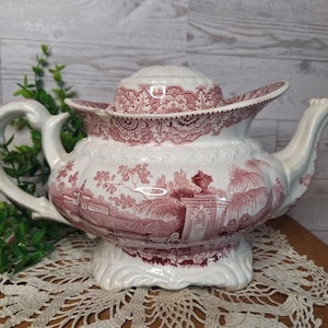May include: A white ceramic teapot featuring a detailed maroon transferware pattern of a landscape with trees, buildings, and a pedestal urn. The teapot has a curved handle, a spout, and a matching lid with a floral border design.