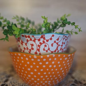 May include: Two decorative ceramic bowls stacked on top of each other. The bottom bowl is orange with white polka dots. The top bowl is white with a red floral design and a blue rim. Green leafy sprigs are placed in the top bowl.