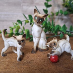 May include: Three ceramic Siamese cat figurines. One cat is standing, two are in playful poses, one with a red ball. The cats have tan and brown markings, green eyes, and are set against a neutral background with greenery.