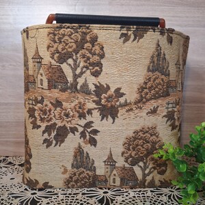 May include: Rectangular storage basket with a tapestry design featuring a landscape scene with trees, buildings, and flowers in shades of brown and beige. The basket has a black handle and a wooden grip.