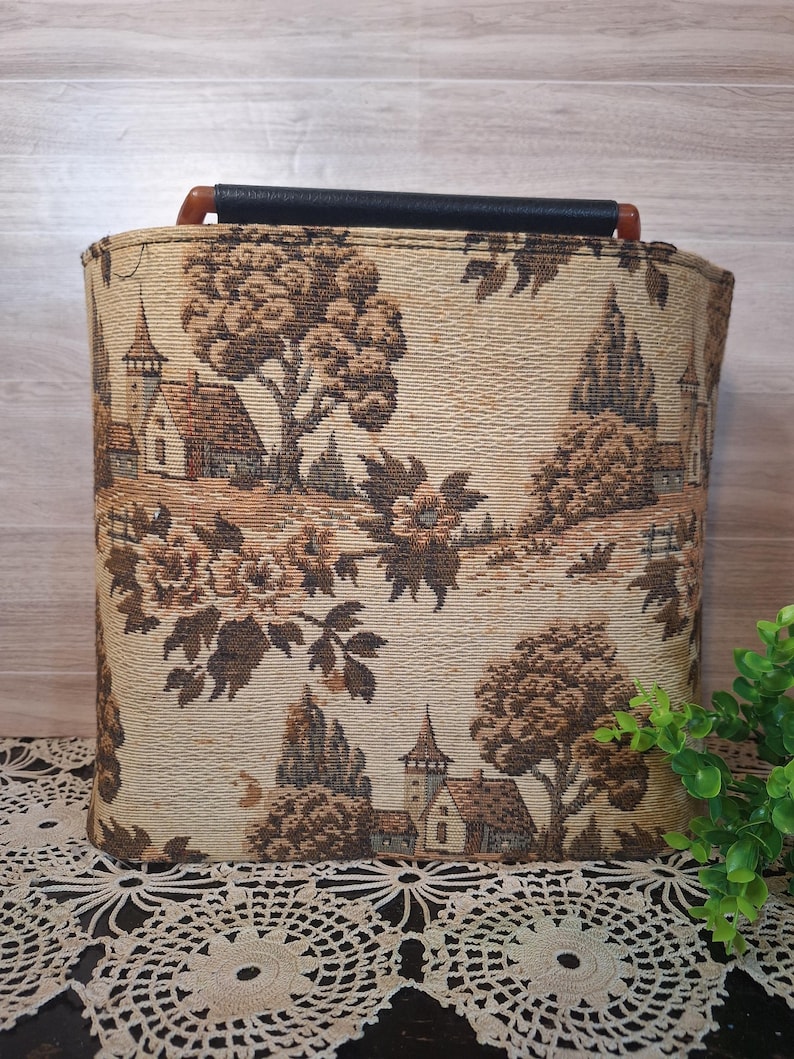May include: Rectangular storage basket with a tapestry design featuring a landscape scene with trees, buildings, and flowers in shades of brown and beige. The basket has a black handle and a wooden grip.