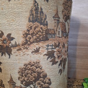 May include: A cylindrical tote bag with a tapestry design. The fabric features a landscape scene with trees, buildings, and a church tower in shades of brown and beige. The bag has a wooden handle.