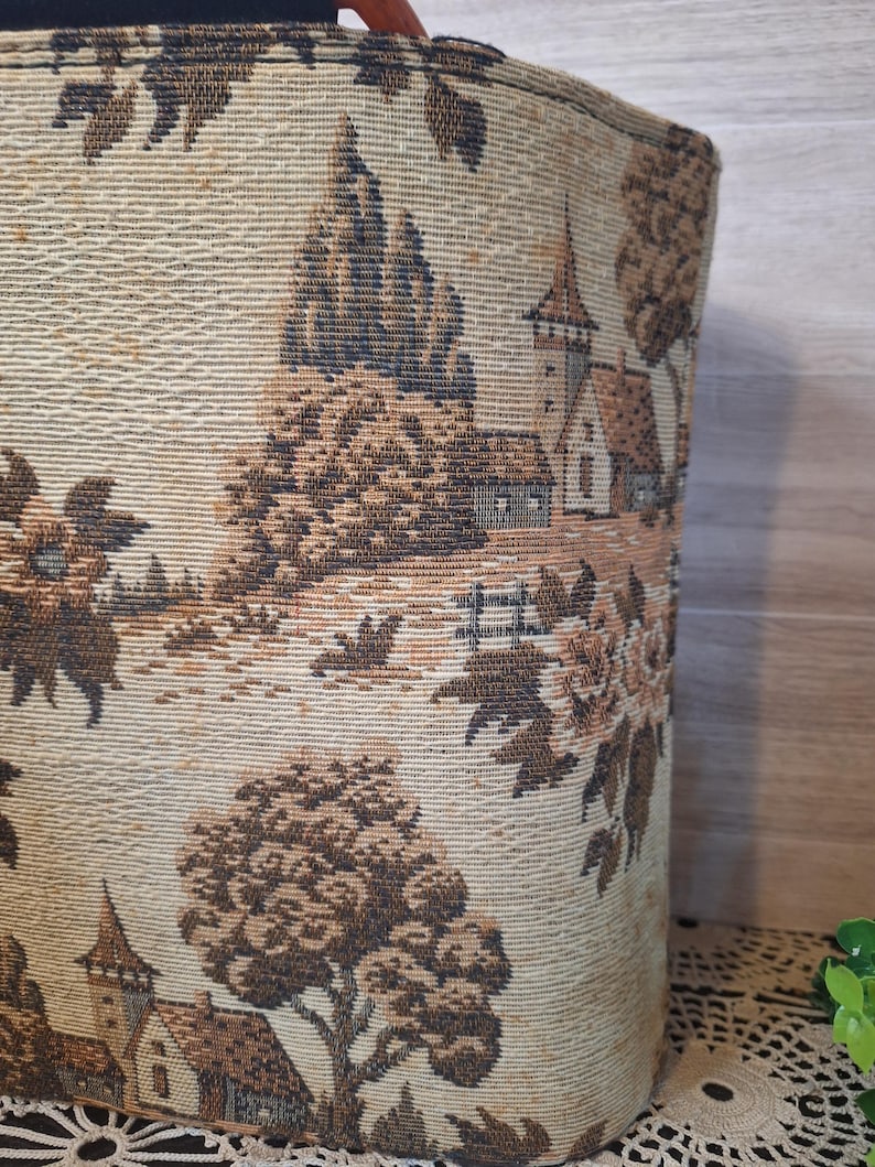 May include: A cylindrical tote bag with a tapestry design. The fabric features a landscape scene with trees, buildings, and a church tower in shades of brown and beige. The bag has a wooden handle.