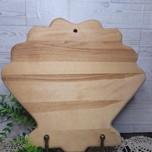 May include: A light-colored wooden cutting board shaped like a seashell with a smooth, natural grain finish. The board features a small circular hole near the top edge for hanging and is supported by a dark metal stand at the base.