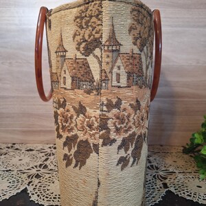 May include: Vintage tapestry handbag with brown wooden handles. The bag features a detailed landscape scene with buildings and trees in shades of brown and beige. The lower portion of the bag has a floral pattern.