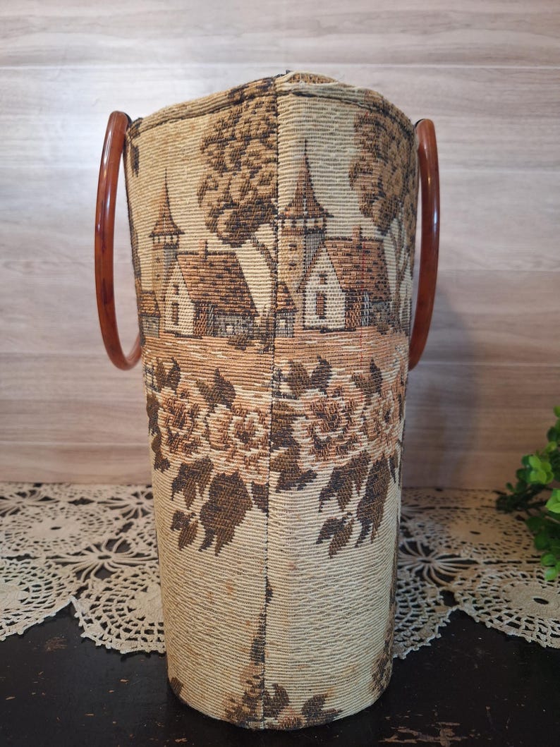 May include: Vintage tapestry handbag with brown wooden handles. The bag features a detailed landscape scene with buildings and trees in shades of brown and beige. The lower portion of the bag has a floral pattern.