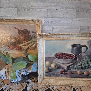 May include: Two framed still life paintings with a rustic, vintage aesthetic. The first painting features a copper pot, wheat, and vegetables. The second painting features a bowl of red berries, grapes, and a pewter pitcher.