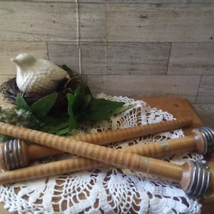 May include: Three wooden spools with metal ends are arranged on a wooden surface, with a white lace doily and a small bird figurine in the background. The spools have a striped design and are likely vintage sewing or craft supplies.