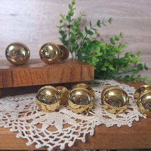May include: A collection of shiny, gold-colored spherical door knobs. The knobs are arranged on a wooden surface with a white lace doily and green foliage in the background. The knobs have a reflective surface.