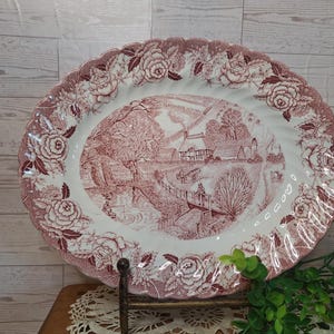May include: An oval ceramic serving platter featuring a deep red transferware pattern. The center depicts a rural landscape with a cottage, a bridge, and a stream. The scalloped rim is decorated with a detailed border of roses and foliage.