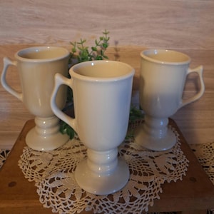 May include: Three off-white ceramic mugs with handles, set on a wooden surface with a lace doily. The mugs have a pedestal base and are filled with a light-colored liquid. Greenery is placed between the mugs.