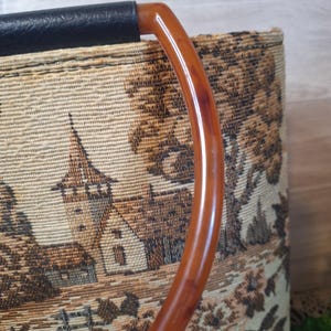 May include: Vintage tapestry handbag with a brown and tan landscape design. The bag features a curved, amber-colored handle and a black leather top. The tapestry depicts a detailed scene of buildings and trees.