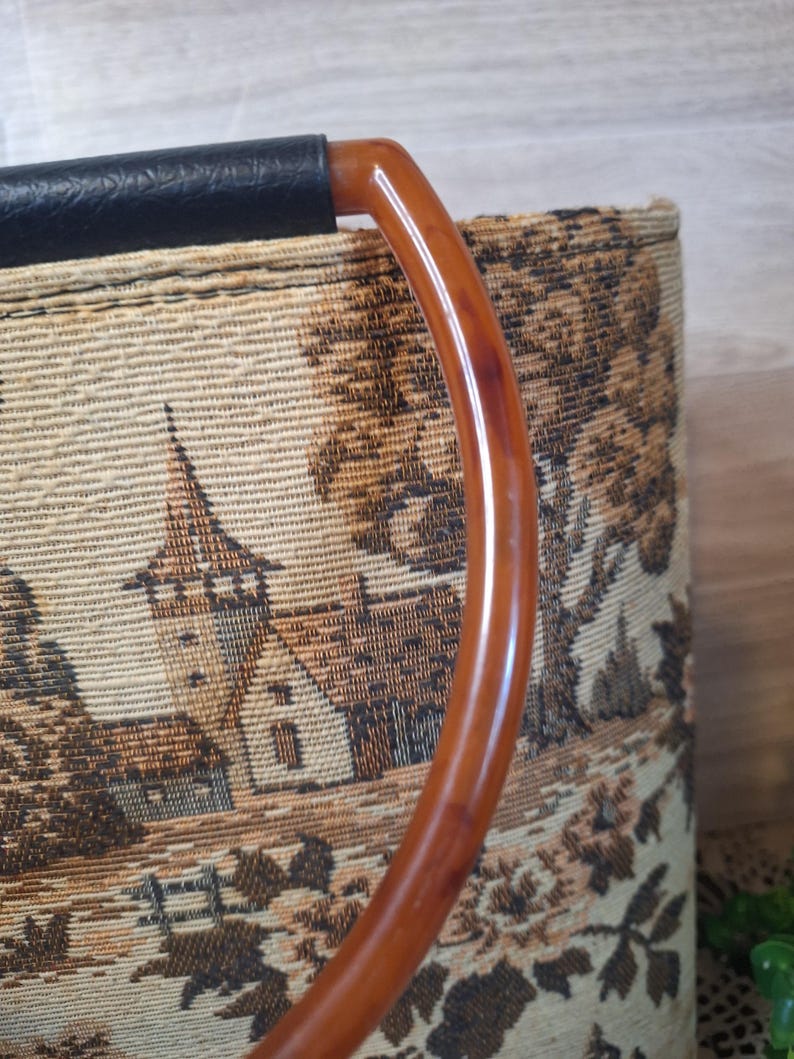 May include: Vintage tapestry handbag with a brown and tan landscape design. The bag features a curved, amber-colored handle and a black leather top. The tapestry depicts a detailed scene of buildings and trees.