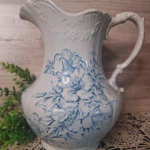 May include: A white ceramic pitcher featuring a blue floral pattern. The vessel has a scalloped rim, an ornate handle, and embossed detailing around the top edge. The body is rounded with a wide base and a prominent pouring spout at the top.