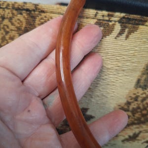 May include: A curved, brown handle with a glossy finish, attached to a black leather section. The handle is held against a patterned fabric background with tan and brown tones.