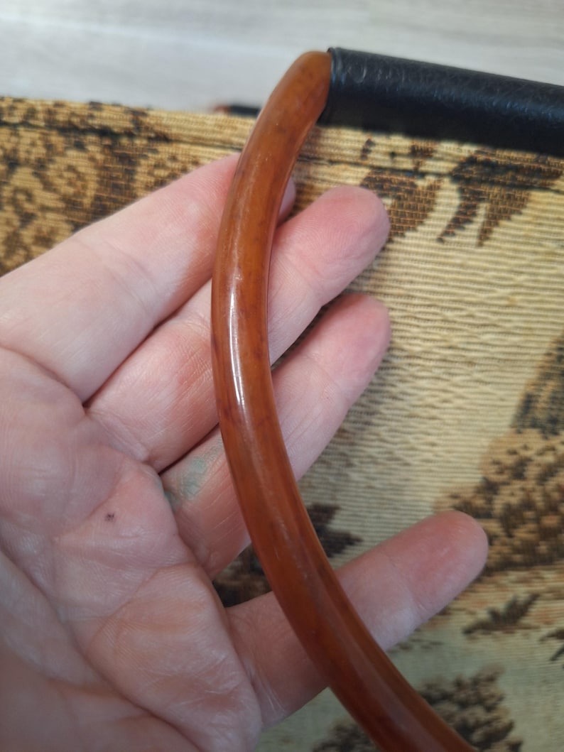 May include: A curved, brown handle with a glossy finish, attached to a black leather section. The handle is held against a patterned fabric background with tan and brown tones.