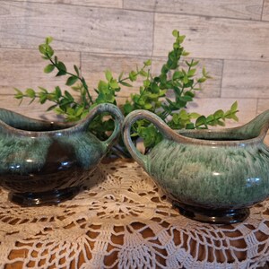 May include: Two green and black ceramic creamer pitchers with a glaze finish. The pitchers have a rounded body and a spout. They are sitting on a crocheted doily.