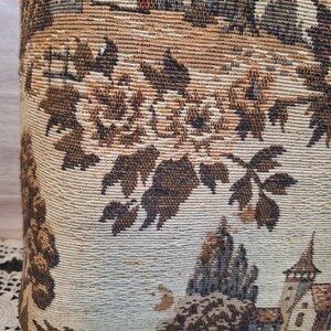 May include: Close-up of a fabric with a detailed tapestry design. The tapestry features a landscape scene with buildings, trees, and foliage in shades of brown, beige, and tan. The fabric has a woven texture.