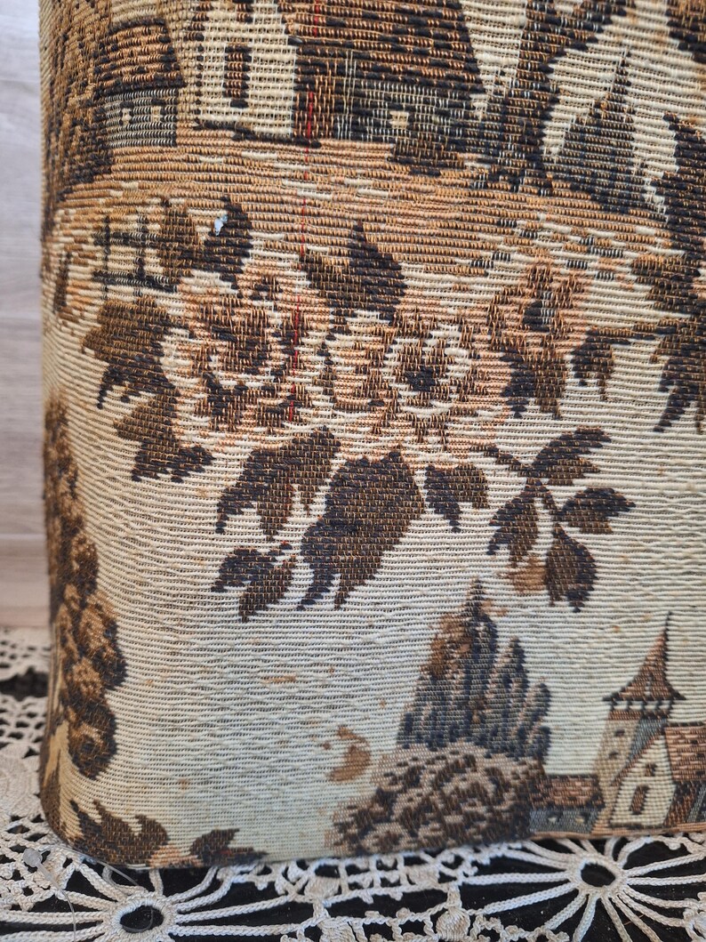 May include: Close-up of a fabric with a detailed tapestry design. The tapestry features a landscape scene with buildings, trees, and foliage in shades of brown, beige, and tan. The fabric has a woven texture.