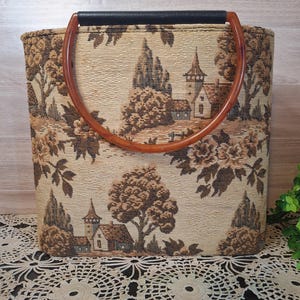 May include: A rectangular tote bag with a tapestry design featuring a landscape scene in shades of brown and beige. The bag has a black handle and brown, arched handles. The bag is sitting on a white lace doily.