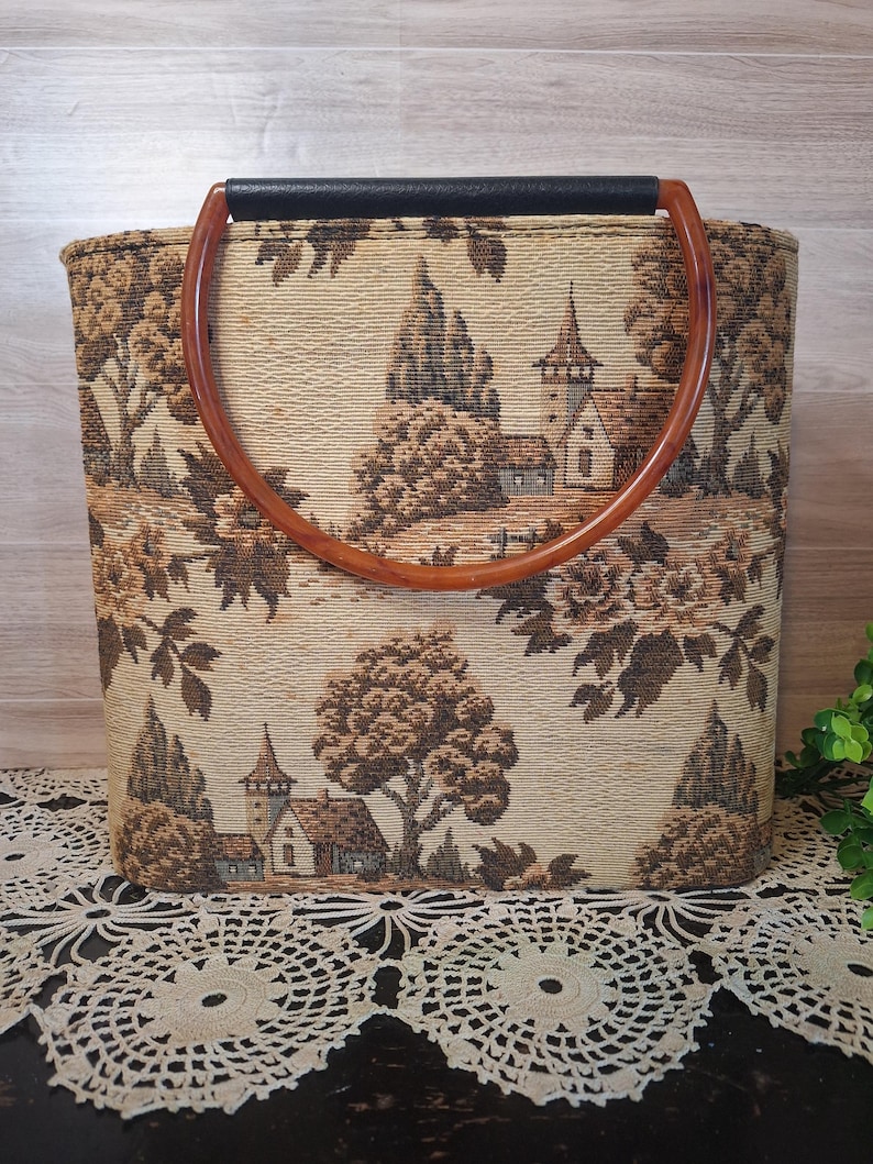 May include: A rectangular tote bag with a tapestry design featuring a landscape scene in shades of brown and beige. The bag has a black handle and brown, arched handles. The bag is sitting on a white lace doily.