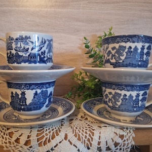 May include: Four blue and white ceramic teacups and saucers. Each cup and saucer features a detailed landscape design. The cups are stacked in pairs, with the saucers beneath. The set is displayed on a wooden surface.