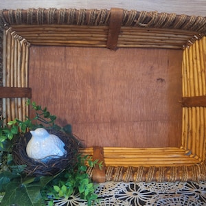 May include: A rectangular, rustic basket with a wooden base and twig frame. A white ceramic bird sits in a nest with green foliage. The basket is trimmed with brown leather straps.