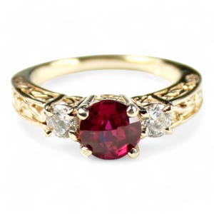 May include: A gold ring with a large, round, red gemstone in the center, flanked by two smaller, clear gemstones. The band features an ornate, engraved design. A classic piece of jewelry.