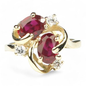 May include: A gold ring with two oval-shaped red gemstones and smaller white gemstones. The ring has a delicate, swirling design.