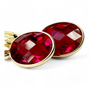 May include: A pair of gold earrings with large, faceted, oval-shaped red gemstones. The gemstones are set in a gold bezel, and the earrings are displayed against a white background. The earrings have a classic, elegant design.
