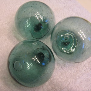 May include: Three green glass fishing floats, each with a small hole at the top. The floats are round and have a slightly textured surface.