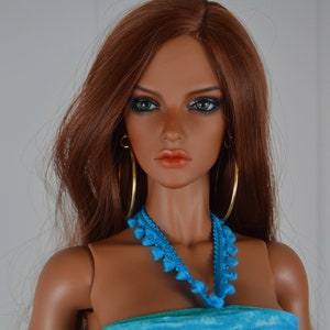 May include: A fashion doll with long auburn hair, green eyes, and full lips. She wears gold hoop earrings, a blue necklace, and a strapless turquoise dress. The doll has articulated joints.