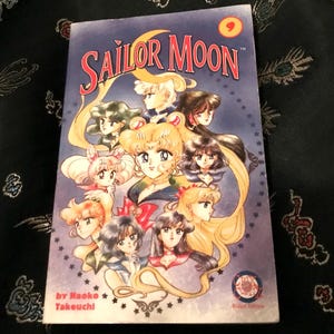May include: Sailor Moon manga volume 9. The cover features a group of anime characters with long hair and colorful outfits. The title is in red and yellow. The background is a dark fabric with a floral pattern.
