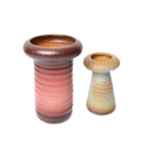 Op de afbeelding: Twee keramische vazen. De grotere vaas is dieprood-bruin met een verloop naar een lichter rood, met een geribbelde textuur. De kleinere vaas is beige en lichtblauw met een glad oppervlak. Beide hebben een cirkelvormige opening.