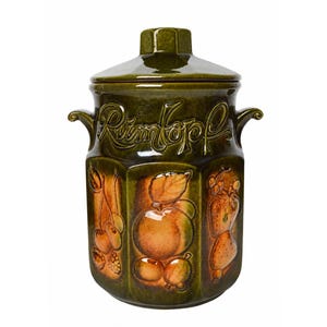 Op de afbeelding: Een donker olijfgroene keramische pot met deksel en twee zijhandvatten. Het woord Rumtopf is in cursief schrift in reliëf aangebracht. De behuizing toont verhoogde, gevormde reliëfontwerpen van diverse vruchten, waaronder kersen, appels en bessen.