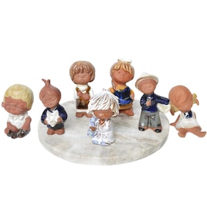 Op de afbeelding: Een set van zeven keramische beeldjes van kinderen, elk met unieke kleding en uitdrukkingen. De beeldjes zijn in een cirkel op een ronde, witte marmeren basis gerangschikt.