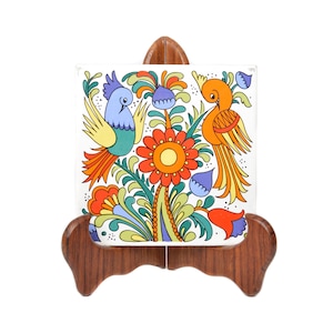 May include: A white ceramic tile with a colourful folk art design featuring two birds and flowers. The tile is on a wooden stand.