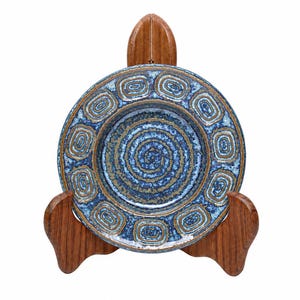 Puede incluir: Un plato redondo de cerámica con un patrón de espiral azul y bronceado en el centro y un borde de formas rectangulares repetidas. El plato se exhibe sobre un soporte de madera de tres puntas con un acabado liso y pulido y veta visible.