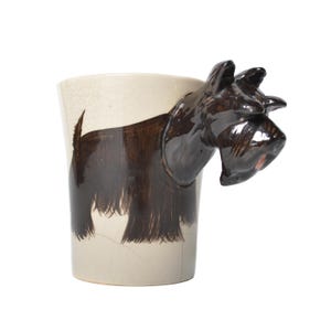 Puede incluir: Una taza de café de cerámica con forma de perro terrier escocés negro. La cabeza del perro es el asa de la taza. La taza está decorada con una imagen en blanco y negro de un terrier escocés.