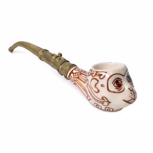Puede incluir: Una pipa de tabaco de cerámica con un diseño de cara de mono. La pipa presenta un cuenco blanco con detalles marrones y un vástago de latón. La cara del mono está detallada con ojos, nariz y boca marrones. El vástago es curvo.