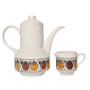 Op de afbeelding: Een witte keramische theepot en roomkan set met een bruin, geel en zwart geometrisch patroon. De theepot heeft een tuit en een handvat. De roomkan heeft een handvat en een tuit.