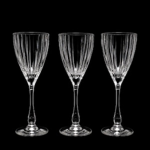 Op de afbeelding: Drie heldere glazen wijnglazen met verticale ribbeldetails langs de kelken. Elk glas heeft een slanke steel met een afgeronde voet en een uitlopende bovenkant, wat een transparant ontwerp toont dat licht reflecteert tegen de donkere achtergrond.