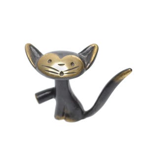 Puede incluir: Pomo de armario decorativo con forma de gato. El pomo tiene un cuerpo negro con cara y orejas doradas. El gato tiene una expresión de sorpresa. La cola se curva hacia arriba. El pomo es probablemente de metal.