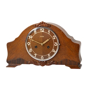Op de afbeelding: Een vintage houten mantelklok. De klok heeft een bruine wijzerplaat met zilverkleurige rand en goudkleurige wijzers. De houten lijst heeft een decoratief ontwerp. De wijzerplaat toont zwarte cijfers.