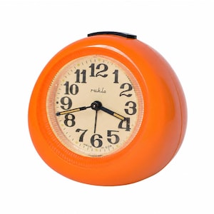 Peut inclure: Un réveil rond orange vif avec un cadran crème, des chiffres arabes noirs et des aiguilles noires. Le nom de marque 'ruhla' est imprimé en écriture noire sur le cadran et le texte 'MADE IN GDR' est visible en bas du cadran.