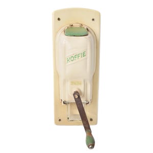 Puede incluir: Molinillo de café vintage de color crema con un mango y detalles verdes. La palabra "KOFFIE" está impresa en verde en la parte delantera. El molinillo tiene una base rectangular y una manivela.