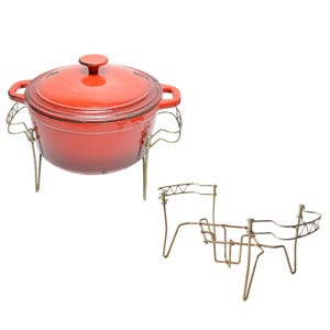 Op de afbeelding: Een rode geëmailleerde gietijzeren pot met een deksel en een gouden metalen standaard. De standaard heeft drie poten en een draadframe.