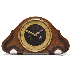 May include: A vintage wooden mantel clock with a dark brown face and gold-colored numerals and hands. The clock has a dark brown wooden frame with black accents and sits on small silver feet. The clock face is circular.
