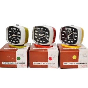 Peut inclure: Trois réveils Bayard vintage avec des cadrans noirs, des bordures blanches et des accents colorés. Chaque horloge est posée sur une boîte assortie avec le nom de la marque et un cercle coloré. Les horloges ont un design rétro.