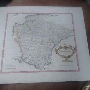 May include: An antique map of Devonshire, England, with a decorative title cartouche in the lower right corner. The map is printed on paper and has a light brown colour.